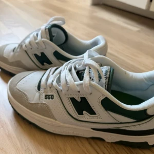 New Balance 550 vita och gröna sneakers - Säljer ett par New Balance 550 sneakers i vitt och mörkgrönt. Skorna har klassisk retrodesign med gröna detaljer, perforerade paneler och mocka framtill. Tjock vit sula och snörning framtill. Perfekta för dig som gillar streetstyle och sportig vibe.