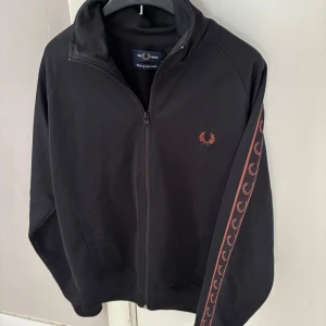Svart Fred Perry zip - Svart ziptröja från Fred Perry med röd broderad logga på bröstet och röda detaljer längs ärmarna. Tröjan har hel dragkedja, ståkrage och två fickor framtill. Materialmix av bomull och polyester, perfekt för en sportig och stilren look.