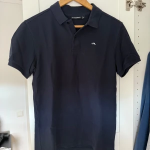 Mörkblå pikétröja från J.Lindeberg - Snygg mörkblå pikétröja från J.Lindeberg med klassisk krage och korta ärmar. Tröjan har en liten vit logotyp på bröstet och knäppning vid halsen. Tillverkad i bomull för skön och luftig känsla. Perfekt för en clean och sportig stil.