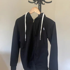 Svart hoodie från Polo Ralph Lauren - Svart hoodie från Polo Ralph Lauren med vit broderad logga på bröstet och vita snören i huvan. Dragkedja framtill och långa ärmar. Perfekt för chill dagar och snygg streetstil.