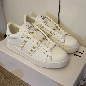 Valentino Rockstud vita sneakers - Snygga vita Valentino Rockstud sneakers i skinn med ikoniska guldiga nitar längs sidorna och på hälen. Klassisk rund tå, platt sula och breda vita skosnören. Perfekta för dig som vill sticka ut med en lyxig och trendig look.