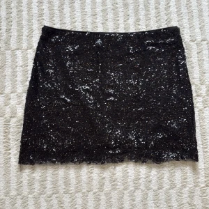 Svart glittrig kortkjol i polyester - Svart kortkjol med glittriga paljetter som ger en riktigt snygg och festlig look. Kjolen är rak i modellen och tillverkad i polyester, vilket gör den både lätt och bekväm. Perfekt för dig som vill sticka ut med lite extra glitter.