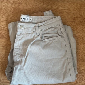 Beiga jeansbyxor från NA-KD - Snygga beiga jeansbyxor från NA-KD reborn med klassisk design och raka ben. Byxorna har bälteshällor, knapp och dragkedja framtill samt en läderpatch med logga bak. Perfekta för en clean och stilren look.