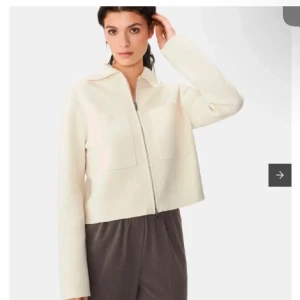 Vit croppad jacka med dragkedja - Snygg vit croppad jacka med dragkedja framtill och två stora fickor på bröstet. Jackan har en klassisk krage och långa ärmar, perfekt för en stilren look. Passar till både jeans och kjol för en trendig outfit.