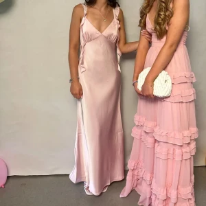 Ljusrosa långklänning i satin  - Superfin ljusrosa långklänning från NA-KD i glansigt satinmaterial. Klänningen har smala axelband, v-ringad urringning och volangdetaljer längs axelbanden. Sitter snyggt med normal passform och ger en elegant look.