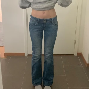 Blå bootcut jeans med låg midja - Säljer ett par blå bootcut jeans med låg midja. Jeansen har dubbla silverknappar, klassiska fem fickor och är i jeanstyg. Säljer då de är för små för mig i midjan