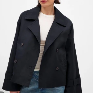 Svart skepparkavaj med bred krage trenchcoat - Säljer en svart skepparkavaj med dubbelknäppning och bred krage. Jackan är croppad med vida ärmar och snygga knappar framtill. Perfekt för dig som gillar en stilren och trendig look. Passar till jeans eller kjol för en cool vibe.