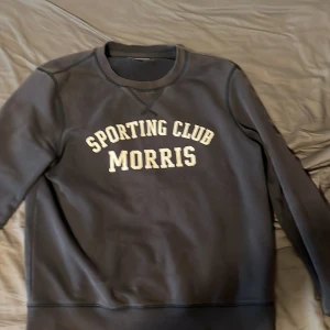 Svart sweatshirt från Morris S - Svart sweatshirt från Morris med trycket 'Sporting Club Morris' i vitt framtill. Klassisk rund halsringning och långa ärmar. Mjuk insida och ribbade muddar vid ärmslut och nederkant. Perfekt för en chill och sportig look.