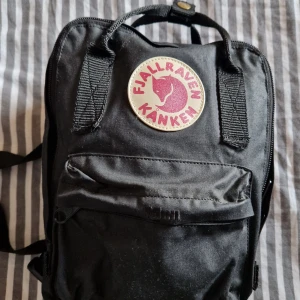 Svart Kånken Mini ryggsäck Fjällräven - Klassisk svart Kånken Mini ryggsäck från Fjällräven med ikonisk rund logga framtill. Väskan har justerbara axelremmar, topphandtag och dragkedja. Tillverkad i slitstark tyg med praktisk ytterficka,sidfickor,t.ex f vattenflaska el paraply och namnlapp på insidan.Oönskad present som inte passar mig perfekt.g.a. liten storlek 