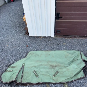 Grönt lippo regntäcke - grönt regntäcke från lippo i slitstark polyester. Utrustad med flera spännen och detaljer i brunt och vitt. och har tydliga etiketter för namn och stall. Mått: 135 cm. 50g.