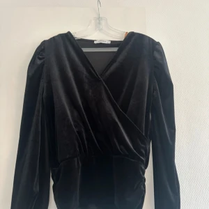 Svart omlottopp i sammet från Chiquelle - Supersnygg svart omlottopp från Chiquelle i mjuk sammet. Toppen har långa ärmar med lätt puff vid axlarna och markerad midja. Perfekt för dig som vill ha en stilren men ändå lyxig look.