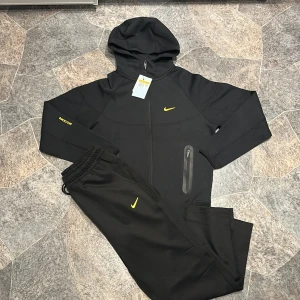 Nike NOCTA svart tracksuit med huva - Svart tracksuit från Nike NOCTA med gul logga på bröstet och ärmen. Setet består av hoodie med dragkedja och huva samt matchande byxor med resår i midjan. Materialet är mjukt och stretchigt, perfekt för chill eller träning. Snygga detaljer med gul NOCTA-tryck och Nike Swoosh.