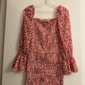 Blommig kortklänning från Parisian - Supersöt kortklänning från Parisian med rött och rosa blommönster på vit botten. Klänningen har långa ärmar med volang vid ärmslut och smockad överdel som ger en snygg passform. Perfekt för dig som gillar romantisk och trendig stil.