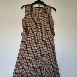 Rutig ärmlös kortklänning med knappar - Säljer en söt ärmlös kortklänning med rutigt mönster i beige, svart och vitt. Klänningen har knappar hela vägen framtill och en lätt A-linjeformad siluett. Den ser ut att vara hemmasydd och har pga slitage en del defekter, främst vid armhålen och knapparna (se bilder för exempel). Skriv gärna vid frågor. 