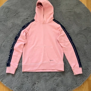 Rosa hoodie från GANT med logga - Säljer en rosa hoodie från GANT med marinblå ränder och vit logga längs ärmarna. Tröjan har huva, magficka och är långärmad. Perfekt för en chill och sportig look. Materialet är mjuk bomull som känns skönt mot huden.
