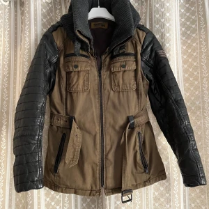 Rockandblue jacka med skinnärmar - Cool jacka från Rockandblue i grönt med svarta, quiltade skinnärmar och snyggt bälte i midjan. Jackan har flera fickor med dragkedja och knappar samt en hög, stickad krage. Perfekt för dig som vill ha en edgy stil med mix av material.