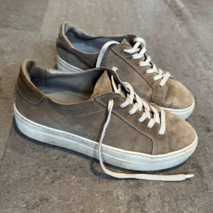 Kaikgröna  mockasneakers med vit sula - Snygga sneakers i beige mocka med vit platt sula och vita snören. Skorna har en rund tå och stilren design som passar till många olika outfits. Perfekt för dig som gillar en clean och enkel look med lite extra edge tack vare mockamaterialet.