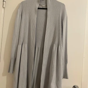 Ljusgrå lång kofta från SCS - Säljer en lång, ljusgrå kofta från SCS med ribbade partier upptill och på ärmarna. Koftan är öppen framtill, har lång ärm och är tillverkad i ett mjukt stickat material. Perfekt att slänga på sig över en t-shirt eller topp för en chill look.