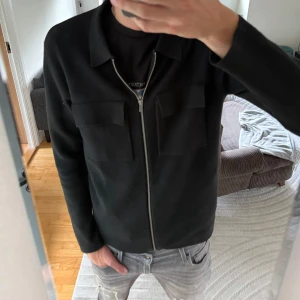 Svart overshirt från Jack & Jones - Stilren svart overshirt från Jack & Jones med två stora bröstfickor och dragkedja framtill. Jackan har klassisk krage och är långärmad, perfekt att slänga på sig över en t-shirt. Materialet känns mjukt och lite strukturerat.