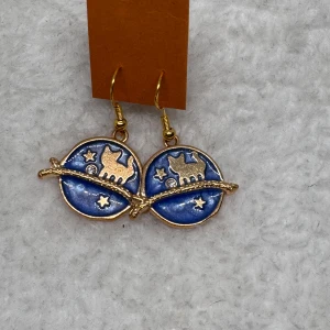 Blå örhängen med kattmotiv - Unika örhängen med rund blå bakgrund och detaljer i guld. Motivet visar en katt, stjärnor och en gren som går över hela smycket. Perfekt för dig som gillar djur och coola detaljer. Hängande modell med krok.