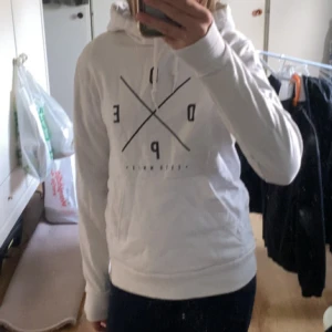Vit hoodie från Dope med tryck - Säljer en vit hoodie från Dope med svart grafiskt tryck på bröstet och klassisk huva med snörning. Tröjan har långa ärmar och ribbade muddar, har lite färg från smink/bus men går nog att få bort i tvätten. 