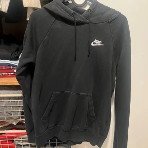 Svart Nike hoodie XS med huva - Svart hoodie från Nike i storlek XS med klassisk logga broderad på bröstet. Tröjan har huva med snörning och en stor känguruficka framtill. Perfekt för en chill och sportig look. Långärmad och i mjukt bomullsmaterial.
