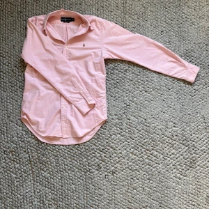 Rosa skjorta från Ralph Lauren  - Klassisk rosa skjorta från Ralph Lauren i bomull med broderad logga på bröstet. Skjortan har lång ärm, knappar framtill och klassisk krage. Passformen är classic fit och materialet känns mjukt och skönt mot huden.
