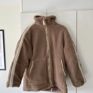 Beige teddyjacka med dragkedja S - Mysig beige teddyjacka med hög krage och hel dragkedja framtill. Jackan har vita detaljer längs ärmarna och två stora fickor. Insidan är quiltad för extra värme och komfort. Perfekt för kalla dagar när du vill vara både varm och snygg.