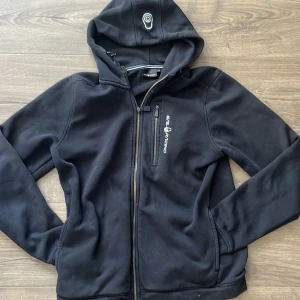 Svart hoodie från Sail Racing med dragkedja - Svart hoodie från Sail Racing med broderad logga på bröstet och huvan. Tröjan har hel dragkedja framtill, bröstficka med dragkedja och mjukt fleecefoder på insidan. Perfekt för kyliga dagar och snygg till jeans eller träningsbyxor.