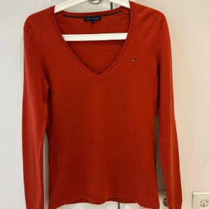 Rostfärgad v-ringad tröja Tommy Hilfiger - Rostorange v-ringad tröja från Tommy Hilfiger med liten broderad logga på bröstet. Tröjan är långärmad och har en klassisk, smal passform. Materialet är mjukt och stickat, perfekt för lager-på-lager under höst och vinter.