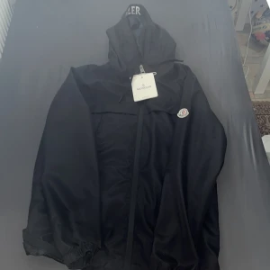 Svart vindjacka från Moncler - Säljer en svart vindjacka från Moncler med huva och dragkedja framtill. Jackan har Moncler-logga på ärmen och tryckt Moncler-text på insidan av huvan. Tillverkad i glansig polyester och har elastiska muddar vid ärmslut. Perfekt för dig som vill ha en stilren och sportig look. Priset kan förhandlas