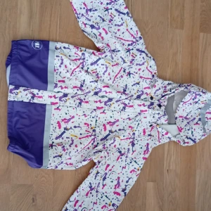 Färgglad regnjacka från PAX med huva - Säljer en vit regnjacka från PAX med färgstänk i lila, rosa och gult. Jackan har lila detaljer nertill, huva och dragkedja framtill. Perfekt för regniga dagar och har en riktigt cool look med sitt unika mönster.