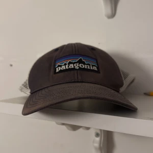 Patagonia keps - Snygg truckerkeps från Patagonia i mörkgrått med vit mesh på sidorna och bak. Framtill finns en broderad Patagonia-logga med bergmotiv i blått, lila och orange. Kepsen har böjd skärm och justerbar snapback. Perfekt för chill och äventyr.