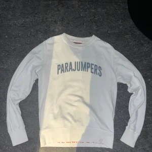 Vit Parajumpers tröja - Vit långärmad fleece-tröja från Parajumpers med tryckt logga i grått framtill och orange detalj på insidan av nederkanten. Tröjan har ribbade muddar och en patch på ena ärmen. Perfekt för dig som gillar clean och sportig stil.