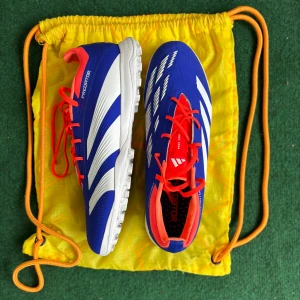 (+påse)Adidas Predator blå fotbollsskor - Säljer ett par Adidas Predator fotbollsskor i blått med vita detaljer och röda snören. Skorna har en låg profil, rund tå och platt sula med dobbar för bra grepp på konstgräs. Ovandelen är i syntetmaterial och insidan har en röd kontrast. Perfekt för dig som vill sticka ut på planen.  Skickar även med en gratis påse som du ser på bild 2. Den är inte så mycket använt, men bara att konstgräs färgen stanna kvar. Den är för Konstgräs,Turf, utomhus skor. 