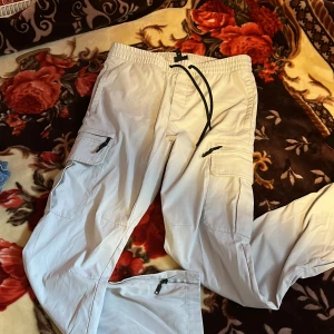 Vita cargopants med dragkedjor - Säljer ett par vita cargopants med elastisk midja och snörning. Byxorna har flera praktiska fickor med dragkedjor och klassiska cargofickor på sidorna. Materialet känns lätt och passar perfekt för en chill och avslappnad stil.