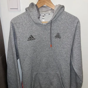 Grå hoodie från Adidas med huva - Snygg grå hoodie från Adidas med klassisk logga på bröstet och justerbar huva med snörning. Tröjan har en stor magficka och är tillverkad i mjukt material som passar perfekt för chill eller träning.
