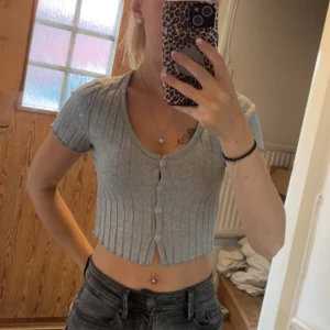 Grå croppad ribbad topp med knappar - Säljer en ljusgrå croppad topp med korta ärmar och v-ringning. Toppen har ribbat material och knappar framtill, vilket ger en snygg och trendig look. Perfekt att matcha med höga jeans eller kjol.