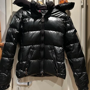 Svart Moncler pufferjacka med huva - Snygg svart pufferjacka från Moncler med glansig finish, huva och dragkedjor både framtill och på fickorna. Klassisk Moncler-logga på ärmen och quiltad design som håller värmen. Perfekt för kalla dagar och riktigt street. size 2