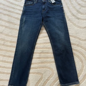 Tommy Hilfiger Bleecker Slim Fit Jeans - Mörkblå jeans från Tommy Hilfiger, modell Bleecker Slim Fit. Byxorna har smal passform, låg midja och klassisk femficksdesign. Tillverkade i stretchigt denim för extra komfort. Snygga detaljer med Hilfiger-logga på bakfickan och läderpatch i midjan. Helt nya med alla tags w33 L30