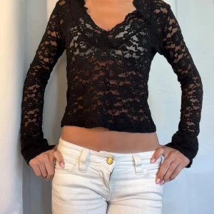 Svart spetsblus med v-ringning - Säljer en svart, transparent spetsblus med v-ringning och långa ärmar. Blusen är croppad och har ett feminint, figurnära snitt. Perfekt för dig som gillar att sticka ut med snygga detaljer och en edgy look.