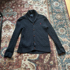 Mörkblå kofta från Polo Ralph Lauren - Snygg mörkblå kofta från Polo Ralph Lauren med kabelstickat mönster och klassisk krage. Tröjan har långa ärmar, knappar framtill och en liten röd logga broderad på bröstet. Perfekt för dig som gillar stilrena och en uppklädd look. Ny pris 3,800 mitt pris 699.