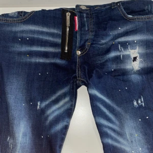 Mörkblå jeans med slitningar och dragkedja - Säljer ett par mörkblå jeans från Dsquared2 med coola slitningar, färgstänk och en stor synlig dragkedja framtill. Jeansen har en klassisk femficksmodell och kontrastsömmar. Perfekta för dig som gillar en edgy streetstil.