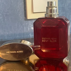 Michael Kors Sexy Ruby parfym 50ml - Michael Kors Sexy Ruby Eau de Parfum i en lyxig, rubinröd glasflaska med guldfärgat lock. Flaskan har en elegant, multifacetterad form som påminner om en ädelsten. Volymen är 50 ml och designen känns både glamorös och modern. Har tappat kroken !! Ser ut som bilder 