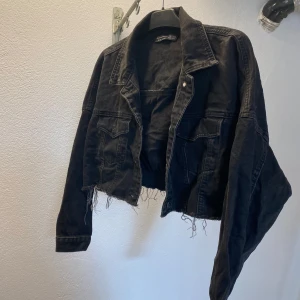 Svart croppad jeansjacka från Dilvin - Svart croppad jeansjacka från Dilvin med rå, fransig nederkant och klassisk krage. Jackan har två bröstfickor med lock och långa ärmar. Perfekt för dig som gillar en edgy och avslappnad stil.