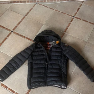 Svart dunjacka från Parajumpers Y-M - Säljer en svart dunjacka från Parajumpers i storlek Y-M. Jackan är superlätt, har huva, dragkedja framtill och två sidofickor med dragkedja. Klassisk Parajumpers-patch på ärmen och quiltad design. Perfekt för kalla dagar när du vill hålla stilen.