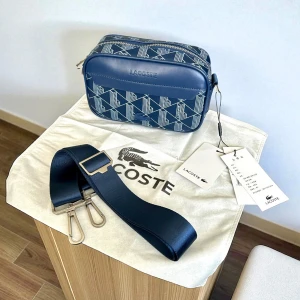 Blå axelväska från Lacoste med mönster - Snygg blå axelväska från Lacoste med geometriskt mönster i vitt. Väskan har justerbar bred axelrem med metallspänne och dragkedja upptill. Tillverkad i slitstark syntet och har diskret Lacoste-logga framtill. Perfekt för att bära det viktigaste med stil.