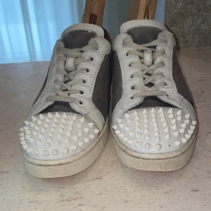 Christian Louboutin sneakers med nitar - Snygga sneakers från Christian Louboutin med klassiska röda sulor. Skorna är grå och vita med coola vita nitar på tån, snörning och detaljer i skinn och canvas. Perfekta för dig som vill sticka ut med en edgy vibe och grich en spik är borta .