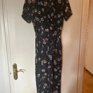 Blommig byxdress från ONLY, marinblå - Snygg marinblå byxdress från ONLY med blommigt mönster i vitt, blått och orange. Byxdressen har omlottliv, kort ärm och knytband i midjan. Vida ben ger en chill och trendig look. Perfekt för dig som vill sticka ut med stil och komfort.