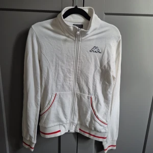 Vit Kappa hoodie med röda detaljer - Vit hoodie från Kappa med dragkedja, broderad logga på bröstet och röda ränder på muddar och fickor. Skön passform och klassisk sportig look, perfekt till chill eller streetwear. Tillverkad i mjuk bomull med långa ärmar.